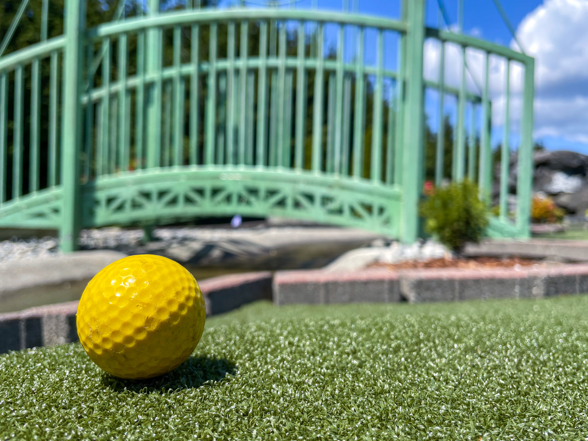 A yellow golfball rests on one of the course greens set against the out-of-focus backdrop of a miniature footbridge designed to resemble the Tacoma Narrows Bridge
